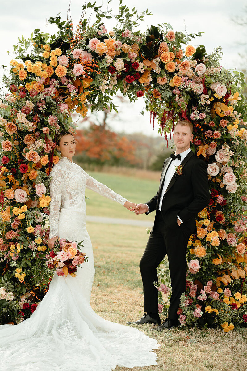 A newlywed couple holding hands and leaning on either side of a floral arch 