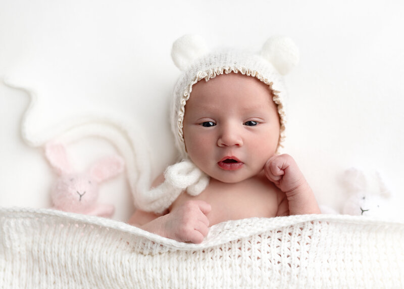 This super sweet baby girl was fully engaged with my camera lense wanting to see and observe everything i did. She is on soft white layers and wearing a bear bonnet and snuggled with our hand knit newborn photo props the pink and white bunny loveys.