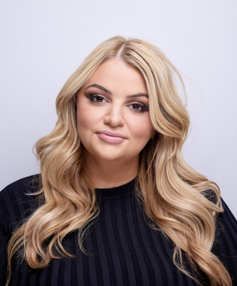 The image features a woman with long, layered hair, wearing lipstick and smiling. The portrait photo highlights her blonde hair and shows her in front of a wall.