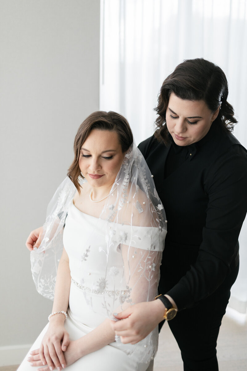 lesbian Palestinian bride in black vest suit placing a veil on her white bride in ivory off the shoulder gown 