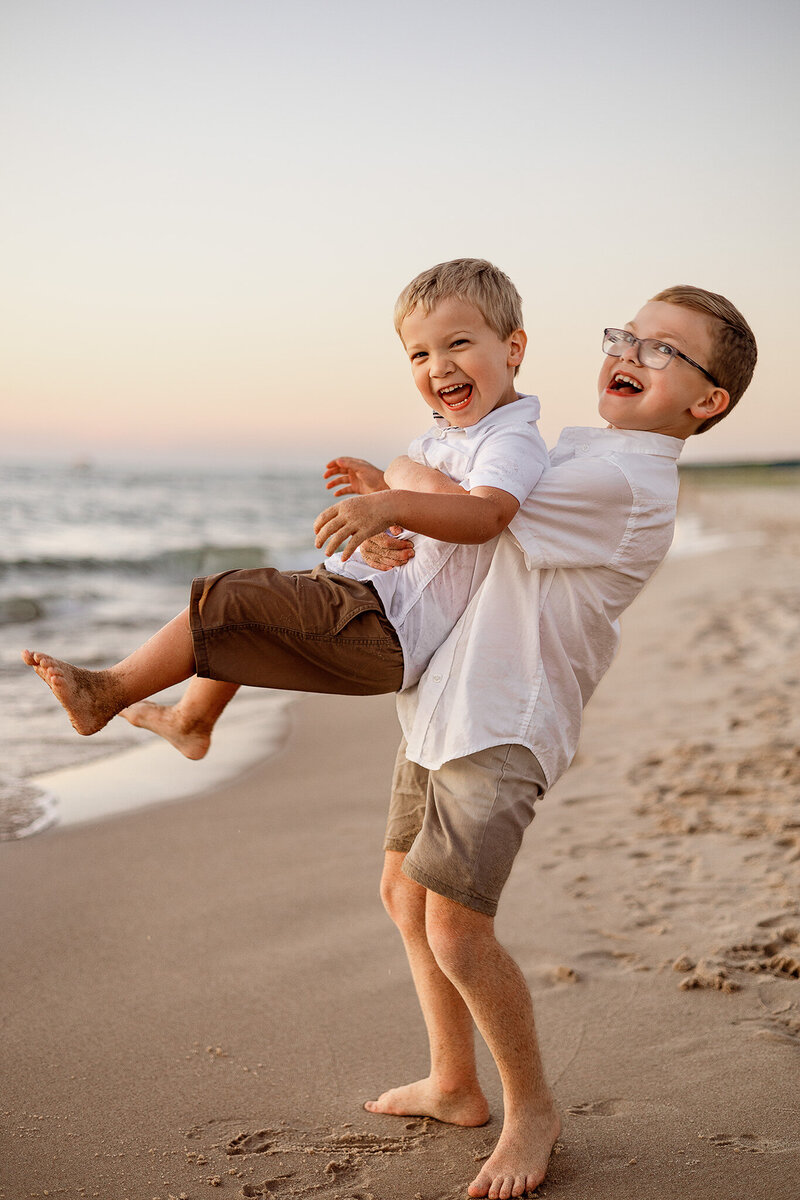 Two young brothers sitting in a field with autumn leaves in West Michigan, capturing a sweet and candid moment as one kisses the other's cheek during a seasonal family photography session.blanket. Captured by Honeywild Photography, a Grand Rapids, MI, luxury newborn photographer specializing in timeless and elegant portraits