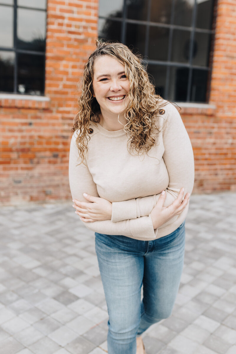 Woman leans towards the camera smiling in front of a brick building. 