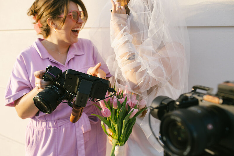 A bride in a wedding dress smiles while holding a camera, ready to document her wedding moments