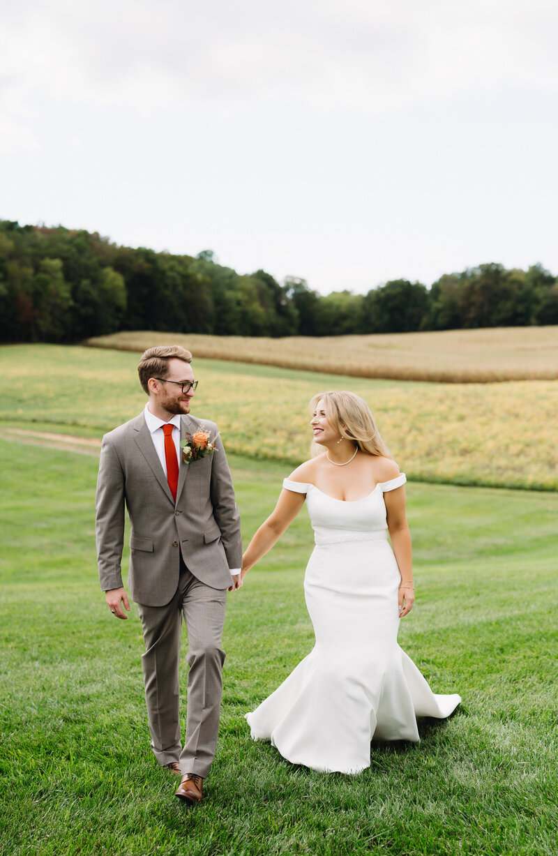 Virginia wedding photographer captures bride and groom holding hands and walking through a bright green field with a treeline in the distance at their Charlottesville wedding venues while they smile to each other for a summer wedding photo