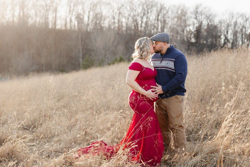 Winter maternity portrait of an expecting couple kissing with mom wearing a beautiful red gown.