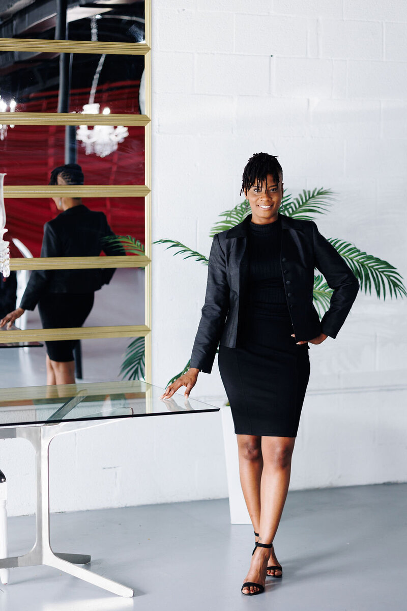 A woman business owner standing near a desk smiling and posing for the camera inside a natural light studio.