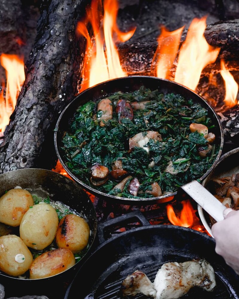 An open campfire with potatoes, greens, and mushrooms cooking in a pan