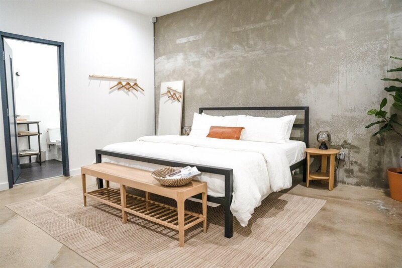 A modern bedroom with a concrete accent wall, a bed with white bedding, a wooden bench at the foot, a small side table, a plant in a terracotta pot, and an open door leading to a bathroom.