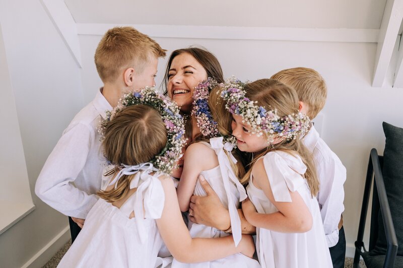 five children in white wiht flower crowns hug bride in akaroa on her wedding day