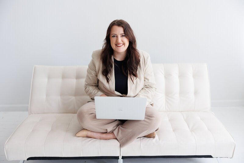 Mariah is sitting on a white couch with her legs crossed and is smiling at the camera. She has her laptop on her lap and is working on a design. 