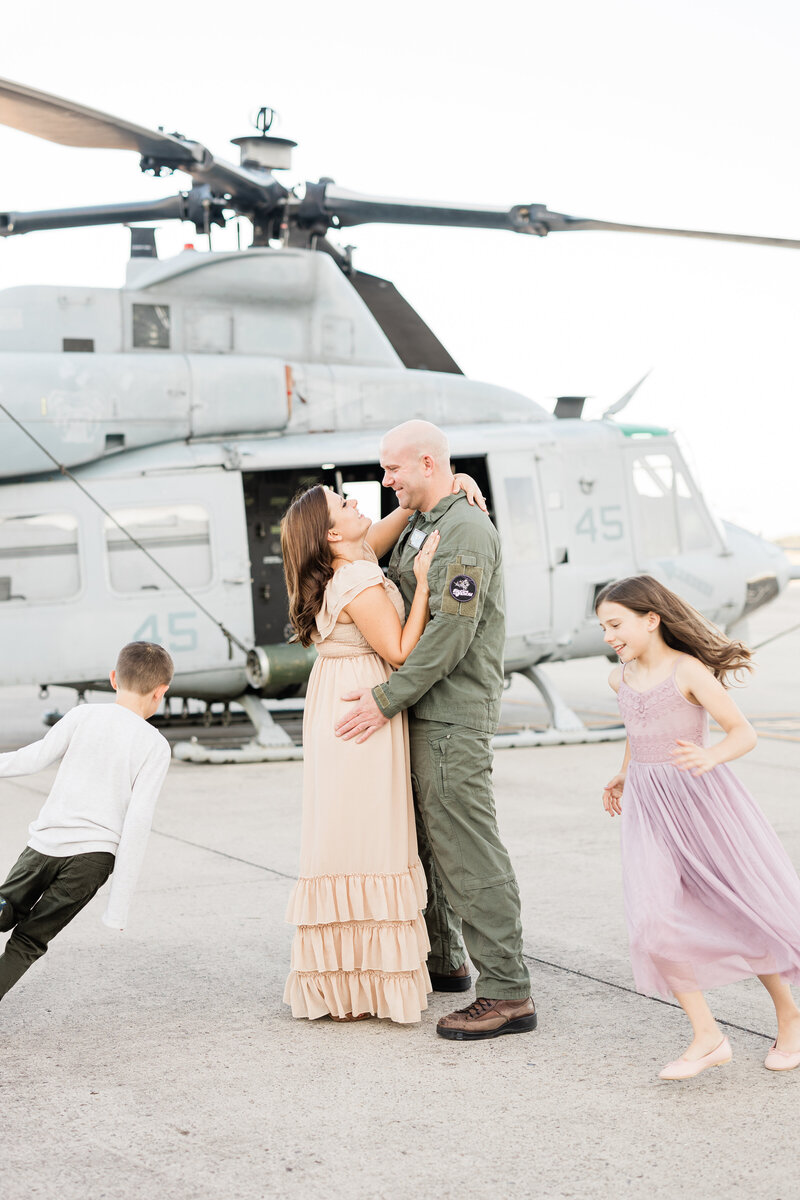 A pilot husband hugs his wife in front of a helicopter while their son and daughter run around them