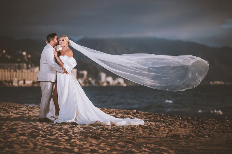 Bride and groom embrace on the beach with dramatic veil and cityscape in background, captured by Houston luxury wedding photographer Giovanni Logrono