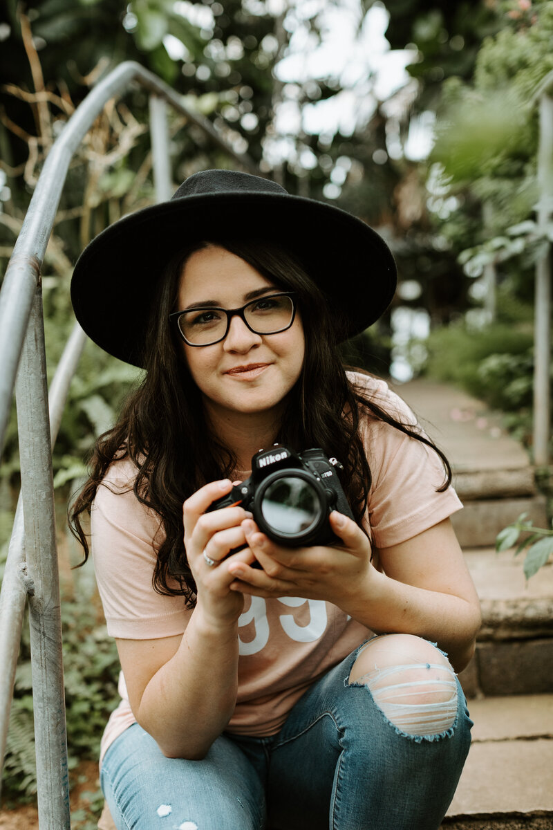 girl with brown hair in ripped jeans and black hat sitting on steps in greenhouse holding a camera