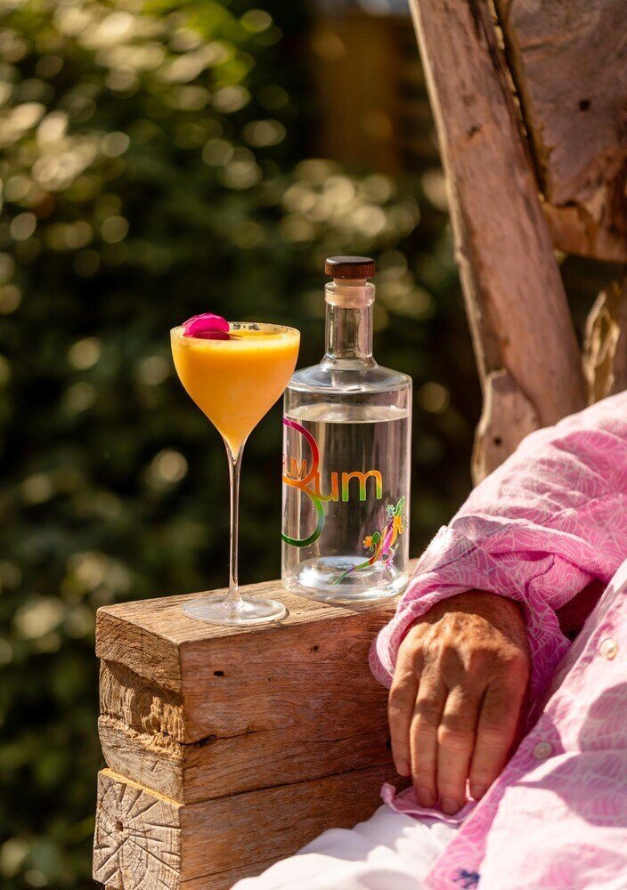 Bottle of Rum Bum rum and a vibrant yellow-orange cocktail garnished with a pink flower, resting on a rustic wooden armrest next to a casually dressed person.