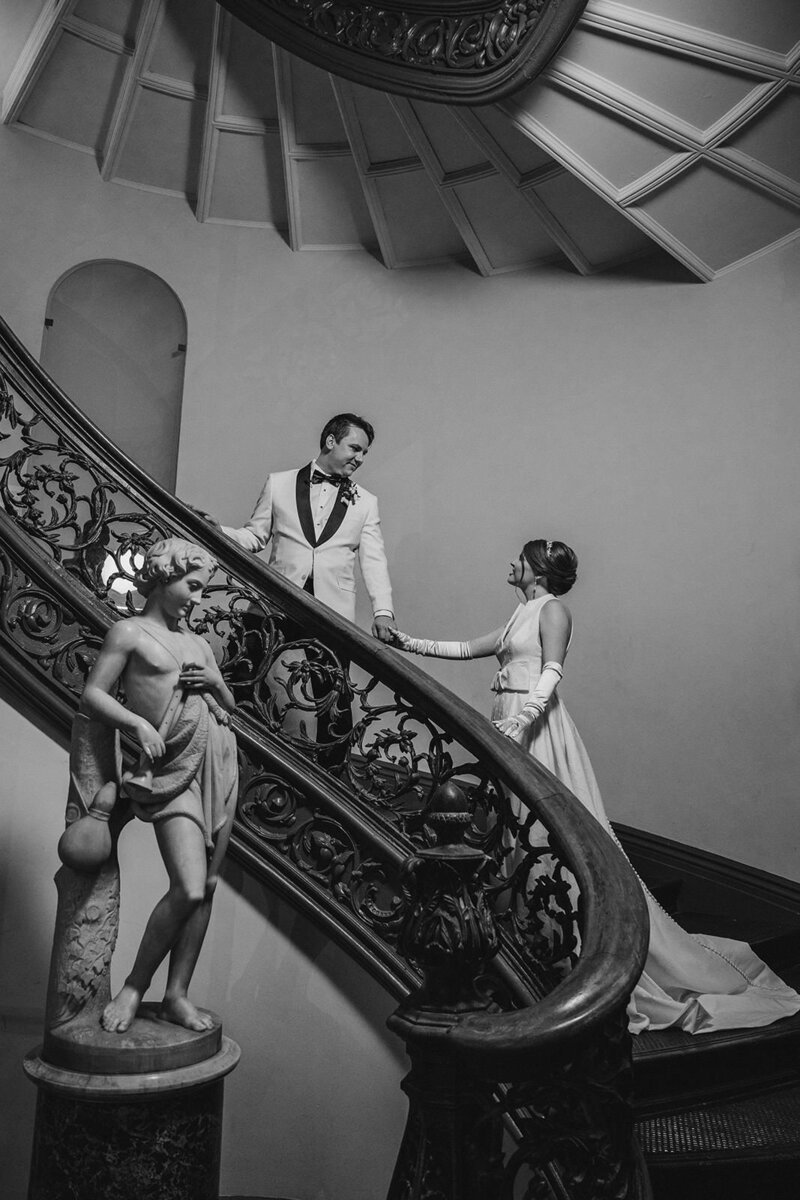 A wedding couple holding hands and walking up a grand staircase 