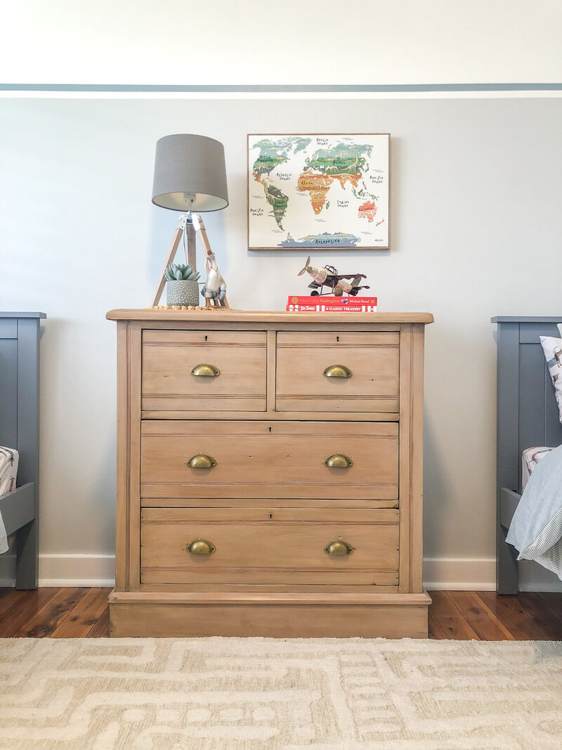 A wooden dresser with a lamp, map, and decor items is flanked by two beds in a cozy bedroom setting. Tailored touches from Custom Interior Design Newcastle enhance the room's charm, bringing bespoke elegance to your living space.