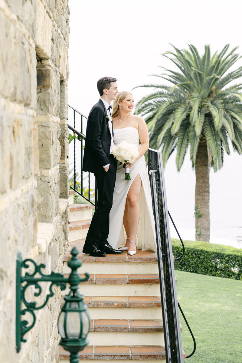 A newlywed bride and groom smiling and standing on stairs overlooking The Lawn at the elegant Bel Air Bay Club in Malibu