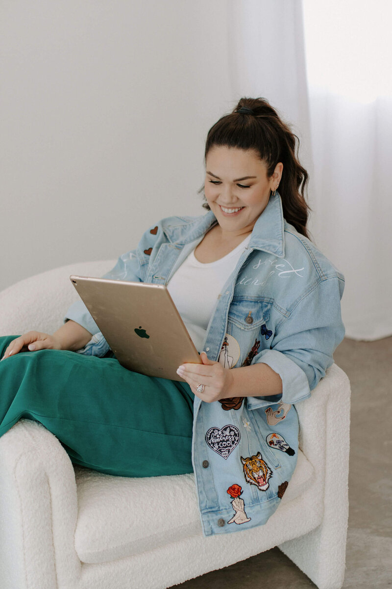 woman with dark hair jean jacket teal pants sitting white chair with ipad