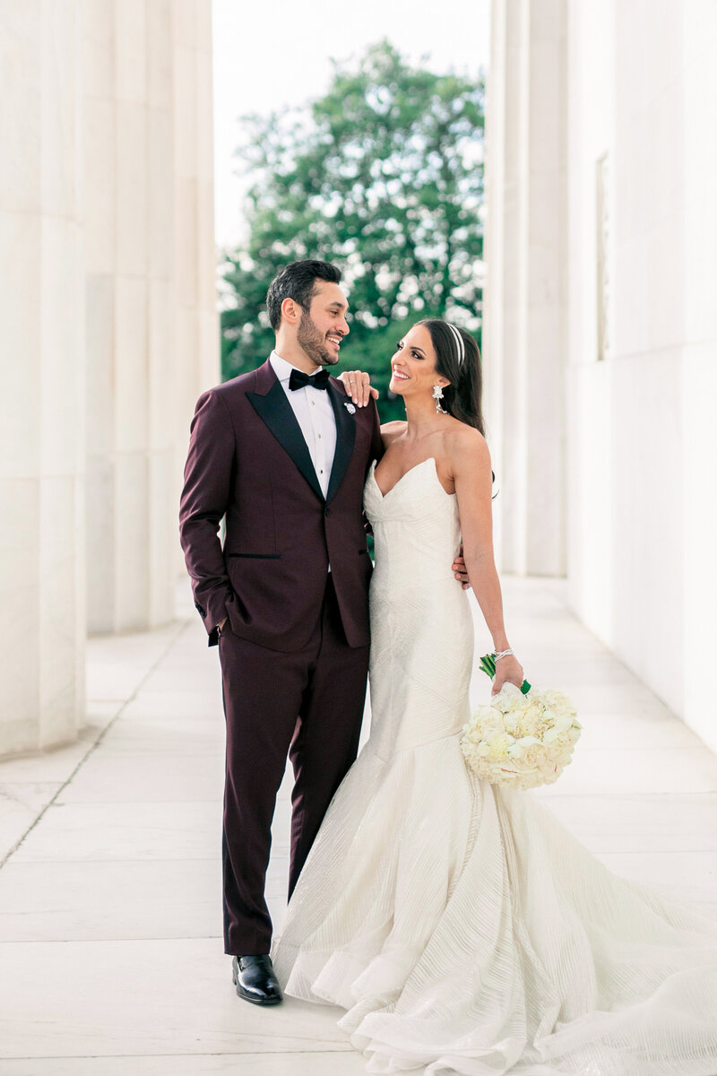 A couple in formal attire poses in an elegant, outdoor setting with a stone pathway and columns. The man wears a maroon tuxedo, and the woman is in a white, strapless wedding gown holding a bouquet of flowers.