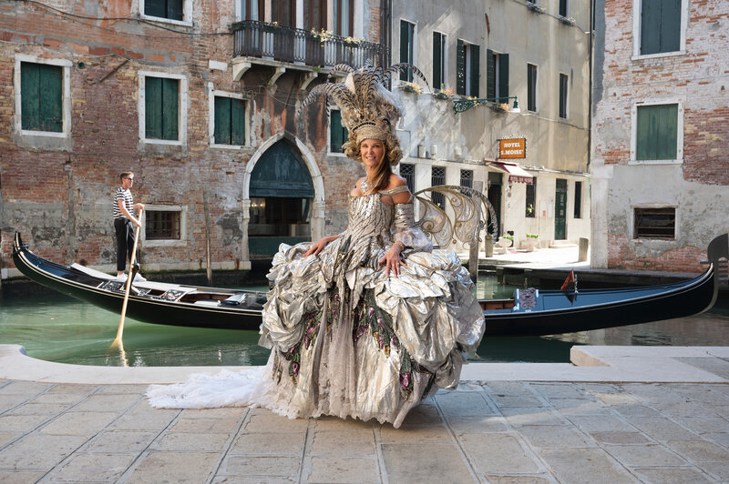 Woman wearing 18th century gown from Venice Carnival designer Antonia Sautter Atelier stands in piazza along canal as gondola passes by. 