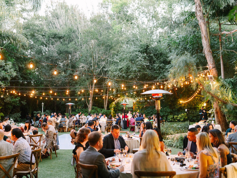 Outdoor wedding reception with guests seated at tables under string lights. Surrounded by lush greenery, the scene is warm and festive, with people enjoying conversations.
