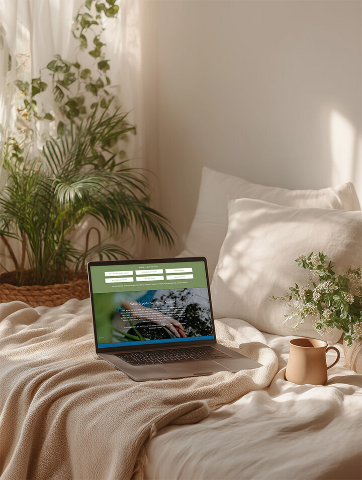 A laptop on a bed next to a plant and a mug, creating a cozy workspace vibe for the author's website.