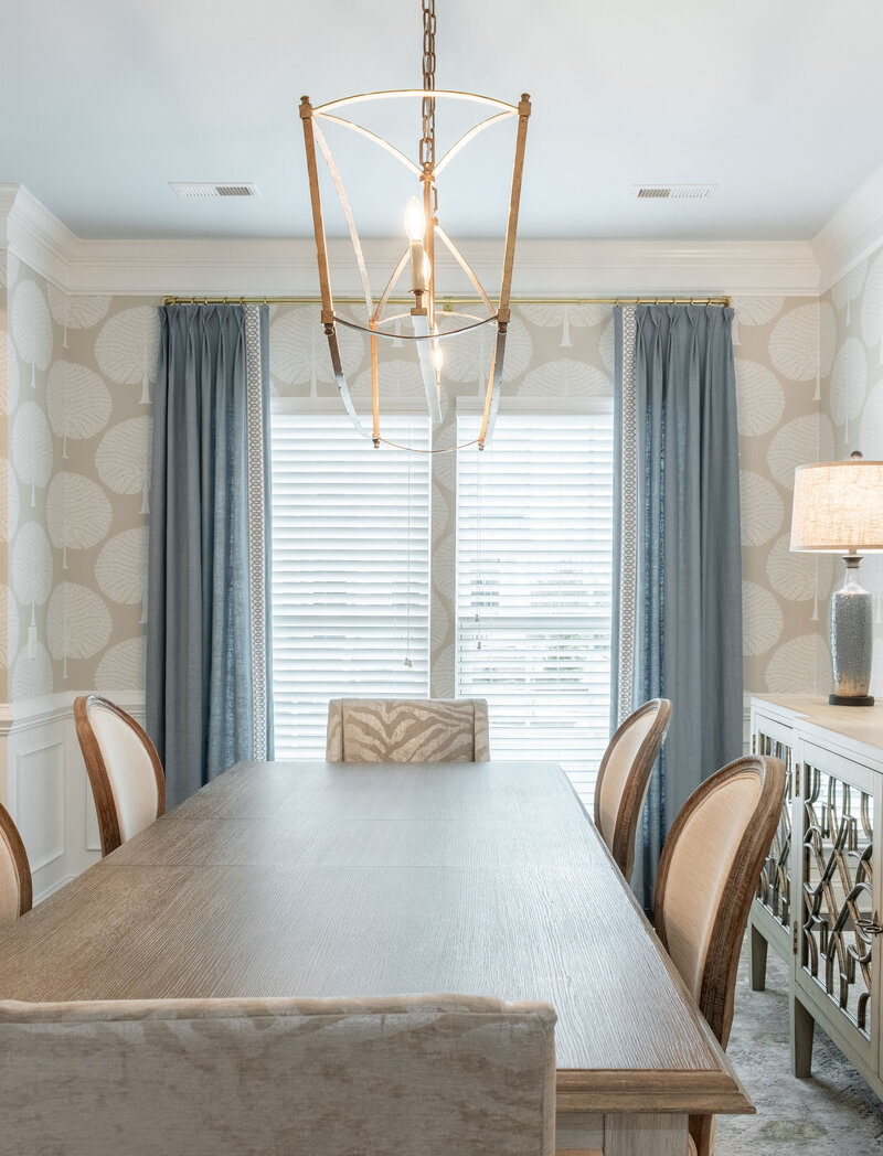 Elegant dining room from The Verlin project by Dwelling Place Interiors, featuring a neutral palette, a wooden dining table with upholstered chairs, and a statement chandelier. Soft patterned wallpaper and mirrored buffet add depth, creating a refined yet inviting space for family dinners.
