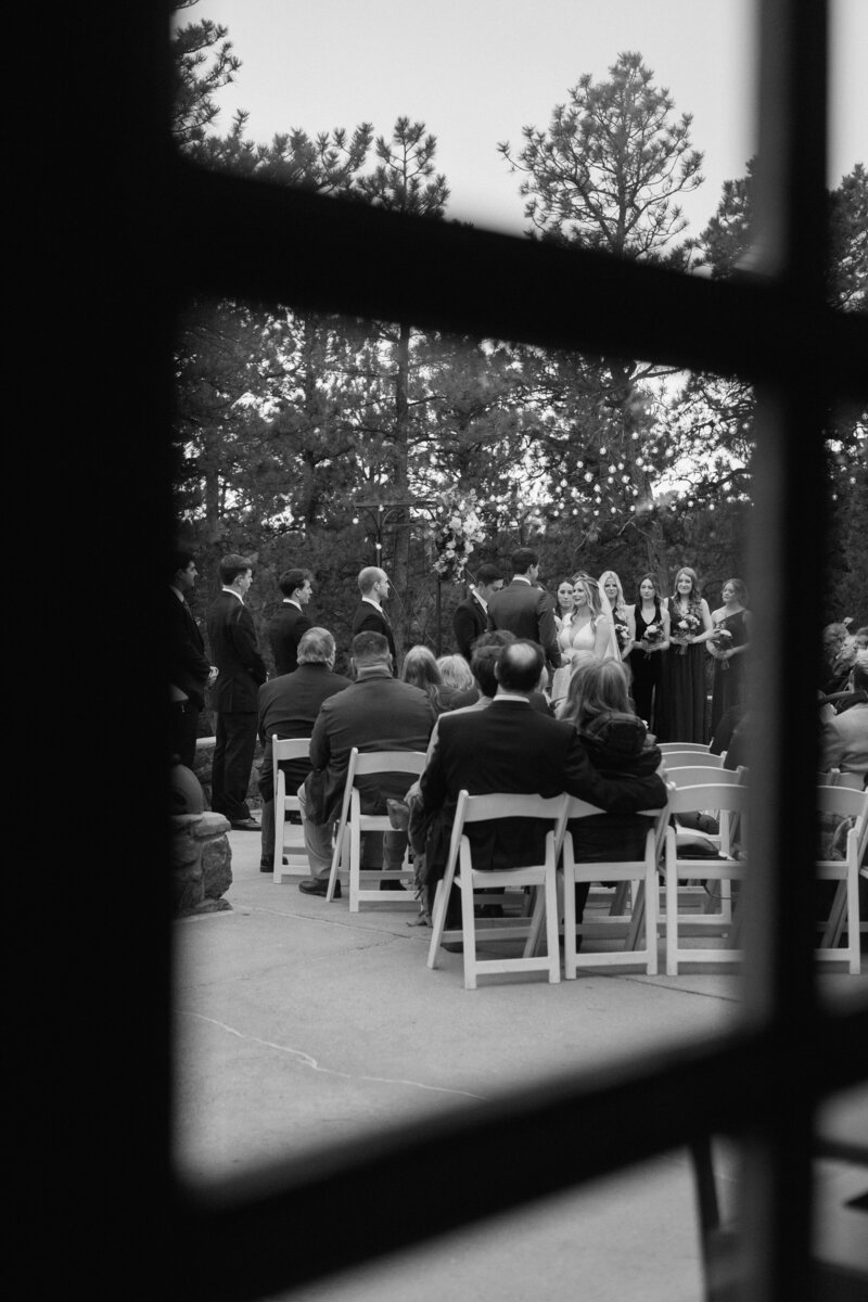 Black-and-white photo of a couple exchanging vows at the altar, captured through a window during a wedding ceremony in a Colorado venue