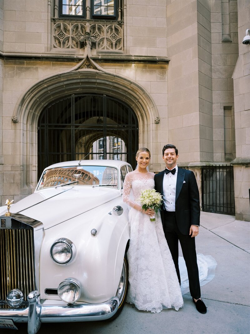 A newlywed couple standing by a classic white vintage car outside a grand building archway, smiling for the camera.