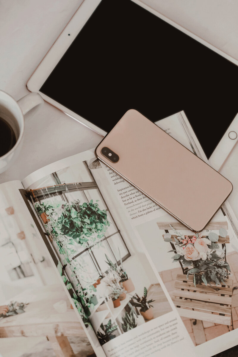 Flat lay of a creative workspace featuring a rose gold iPhone, white iPad, open lifestyle magazine with floral and interior design imagery, and a cup of black coffee on a white marble desk—capturing Sable & Rose’s modern aesthetic and intentional approach to business and everyday inspiration.