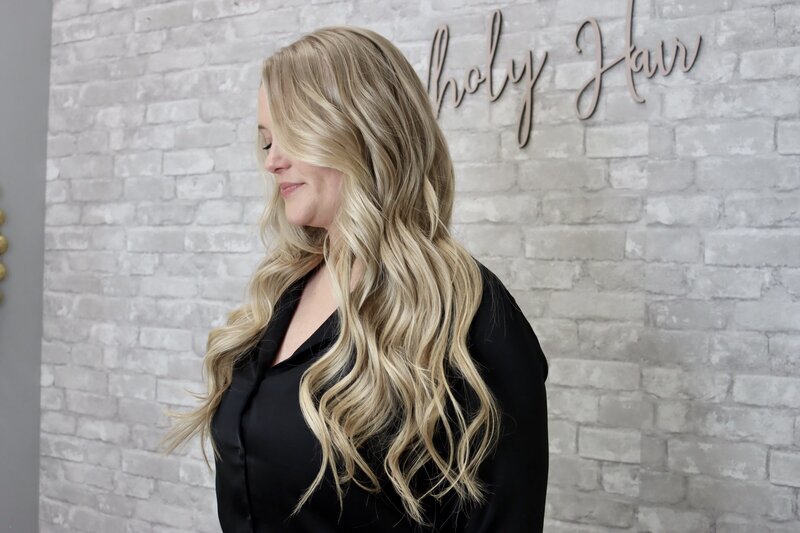 woman with long platinum blonde hair extensions gently smiling with wholy hair sign on the wall