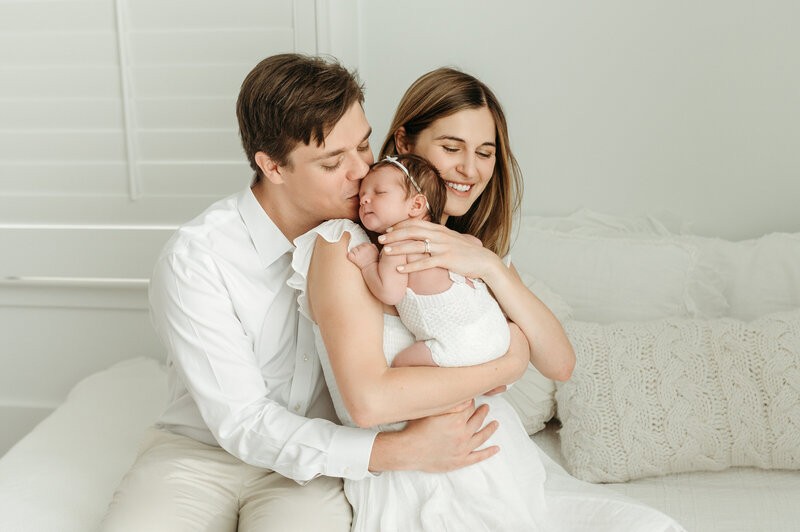 Newborn laying on her mothers shoulder while her dad gives her a kiss. The picture is taken in a studio in Atlanta