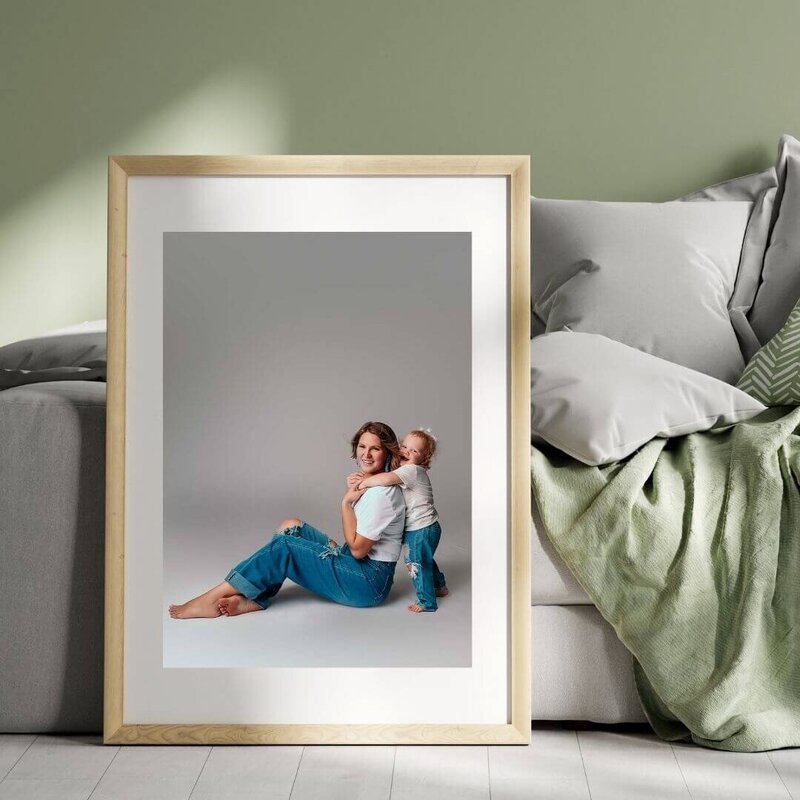 A wall art mock up created by Mobile, AL Family and Motherhood Photographer. The mock up showcases a large-scale print with a white matboard inside a wood frame. The print is on the floorr, propped against a cozy couch with a light sage green blanket which is the same color as the paint on the wall behind the couch. The photo in the mockup print is a mom and toddler daughter duo wearing a matching outfit of blue jeans and t shirt.