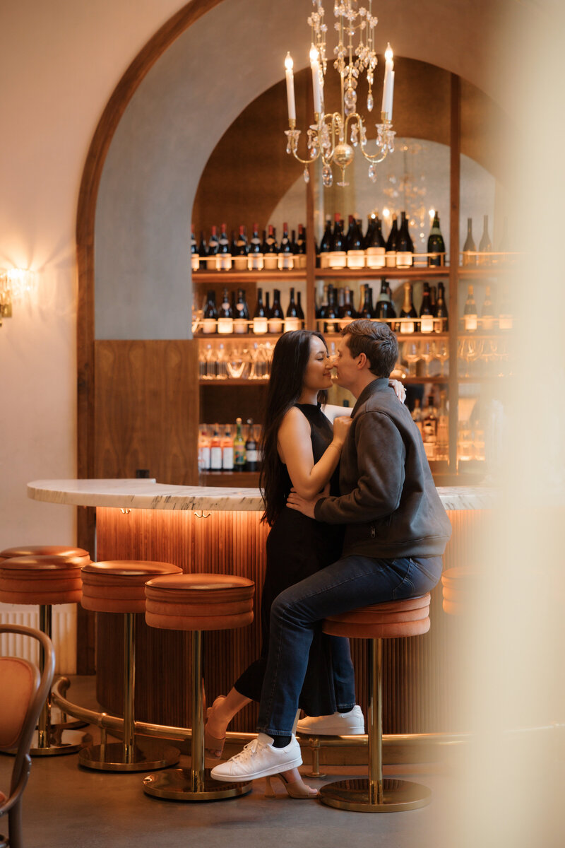 A couple sharing an intimate kiss at the elegant Le Bon Nosh bar in Atlanta, surrounded by warm lighting and wine bottles on display.