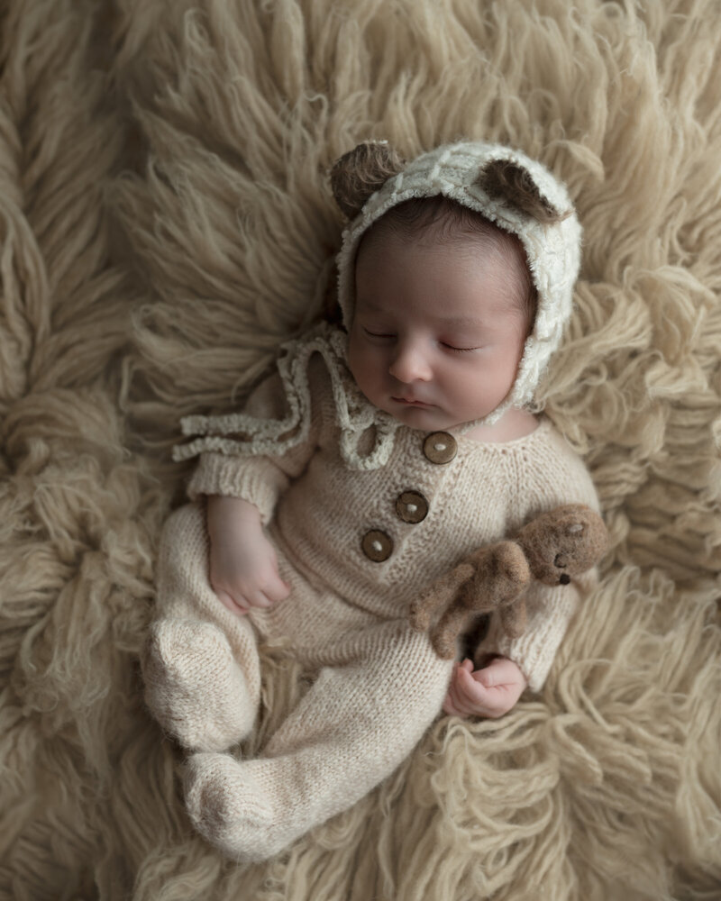 Creamy textures and whimsical bear details surround this baby boy, captured sleeping in a soft knit romper and bonnet on a flokati rug.