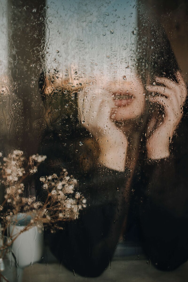 A brunette woman in a black sweater behind a blurry rainy window next to a vase with flowers.