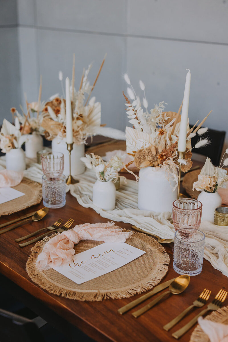 Wedding table styled with neutral tones, dried floral arrangements, gold candlesticks, pink glassware, and woven placemats with knotted blush napkins for a boho-chic reception look.