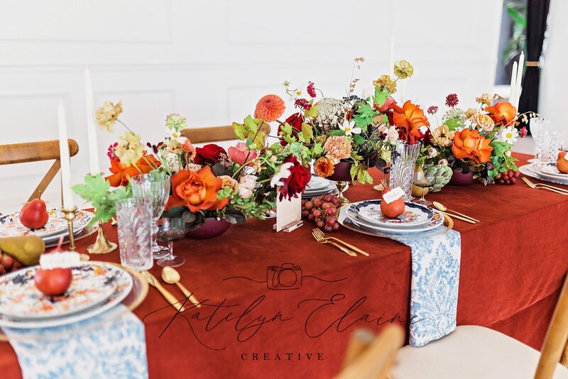 Burnt orange velvet linen and vibrant floral centerpiece arranged on a regular foldable rectangular table with golden wood chairs.