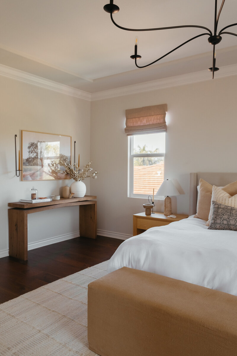Warm and airy San Diego bedroom featuring neutral layered bedding, natural oak nightstand, rustic wood console table with curated styling, and a black sculptural chandelier — a perfect blend of California casual and organic modern design.