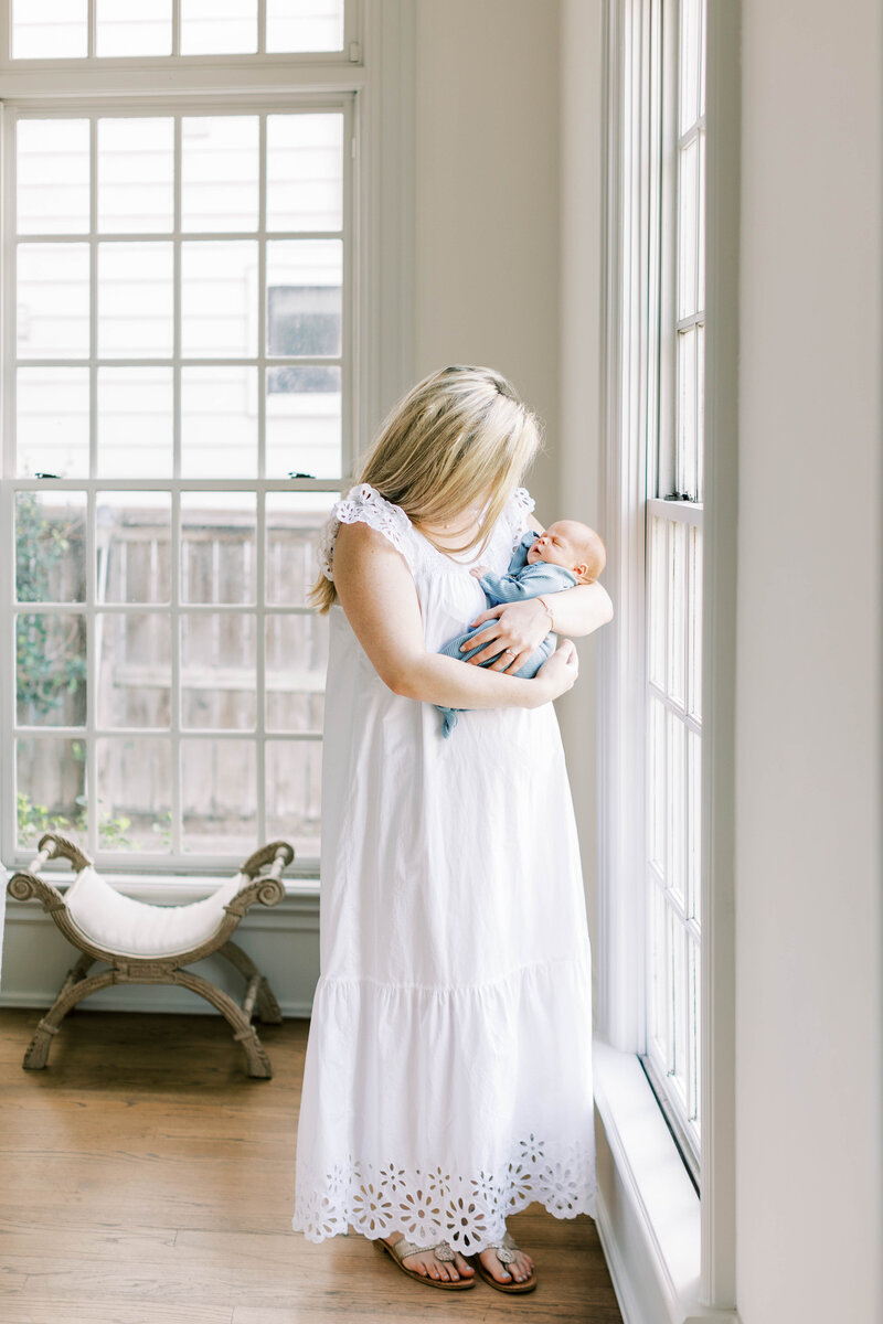 Mom in long white dress holding newborn baby in front of a window