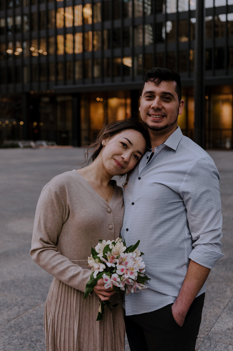 A couple embracing and smiling in an urban setting, with the woman holding a bouquet of flowers.