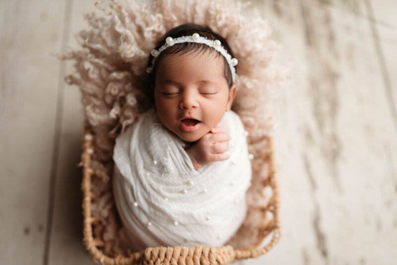 Newborn baby girl swaddled in white swaddle with pearls inside wicker basket and white wooden floor drop