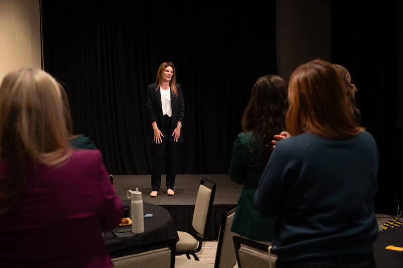 Melanie is wearing a navy suit and cream shirt standing on a stage as the audience claps
