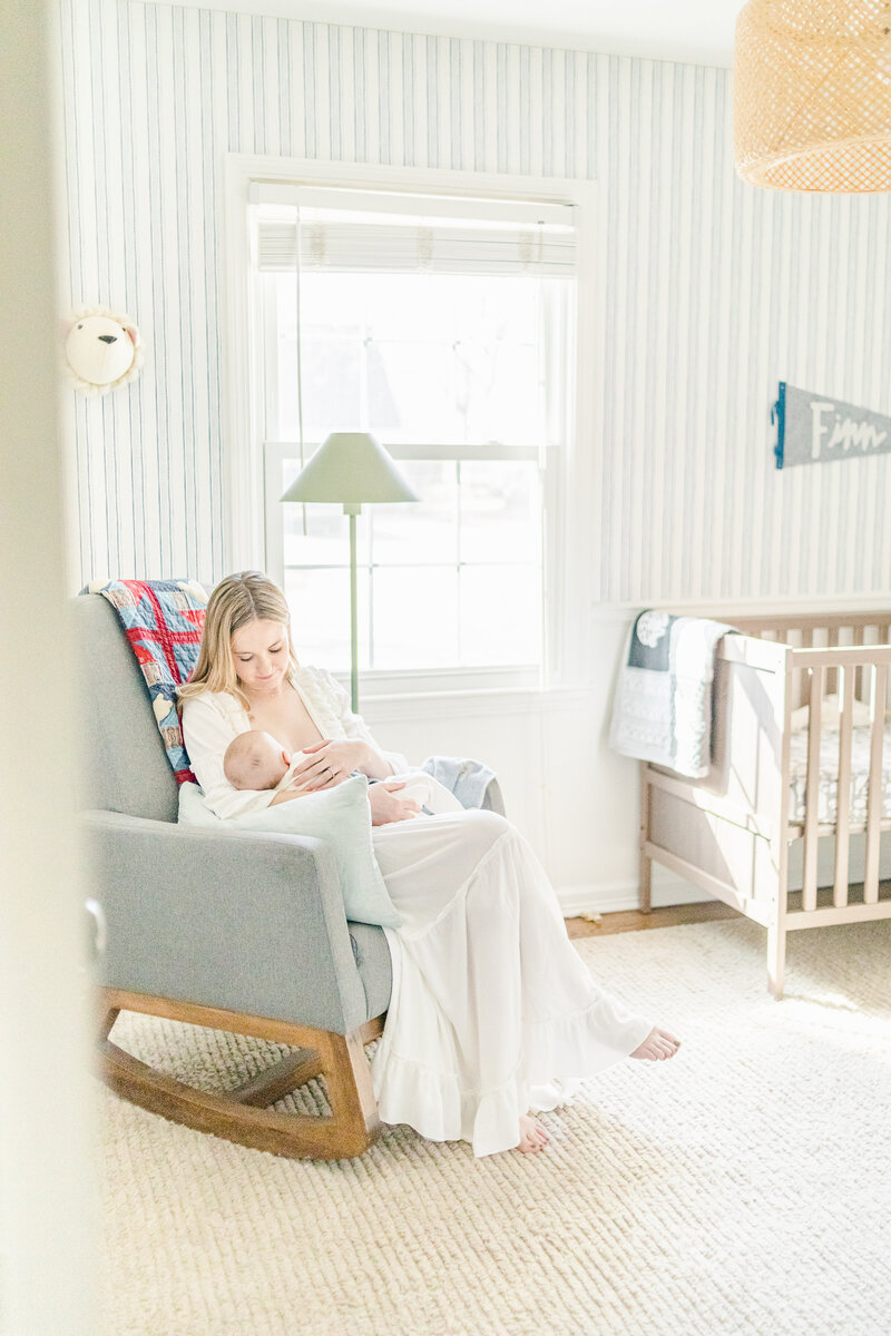 A mother in a long white dress rocks her newborn in a soft-toned nursery filled with natural light, captured by Kansas City newborn photographer Meredith Maag.