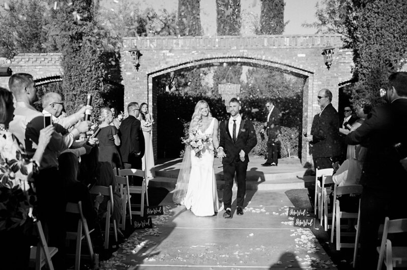 black and white photo of a couple walking down the aisle covered in confetti after becoming husband and wife