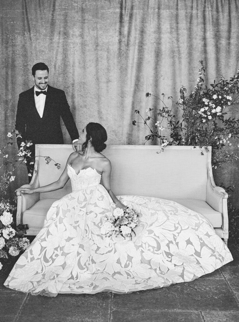 Bride sits on couch in designer gown looking back at groom standing behind couch in Austin, Texas wedding