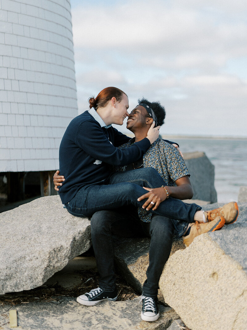 A male gay couple pose on a beach in front of a lighthouse. One partner is a black man wearing a floral shirt and jeans with a bandana around his head. The other partner is a white man with red hair wearing a sweater with collared shirt and jeans and is sitting on his partner's knee. They have their arms wrapped around each other and are smiling with their faces close together.