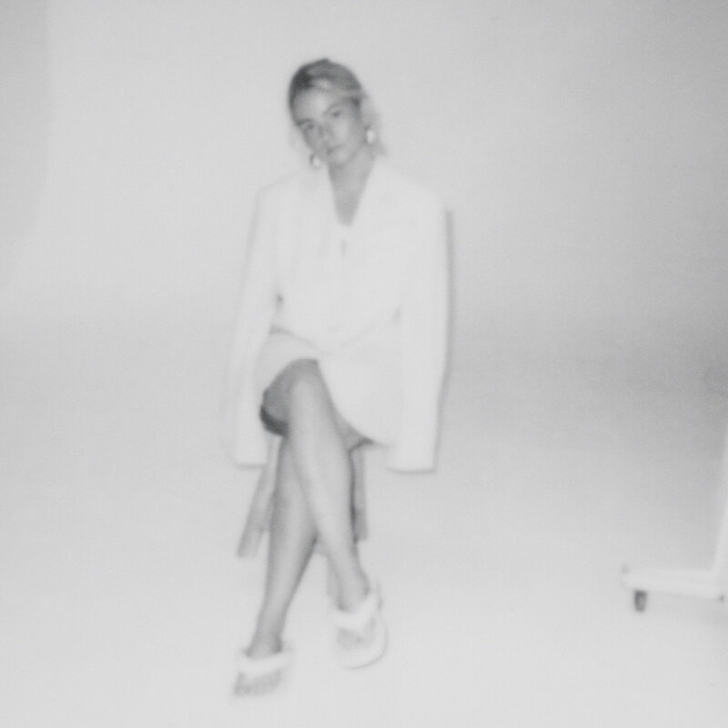 A female wearing a white blazer dress and heeled white sandals sat on a stool in a photo studio