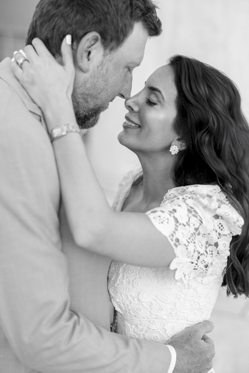 A black and white image of a couple embracing closely. The woman, wearing a white lace dress, holds the mans face. The man is wearing a light-colored suit. Both have eyes closed, appearing intimate and content.
