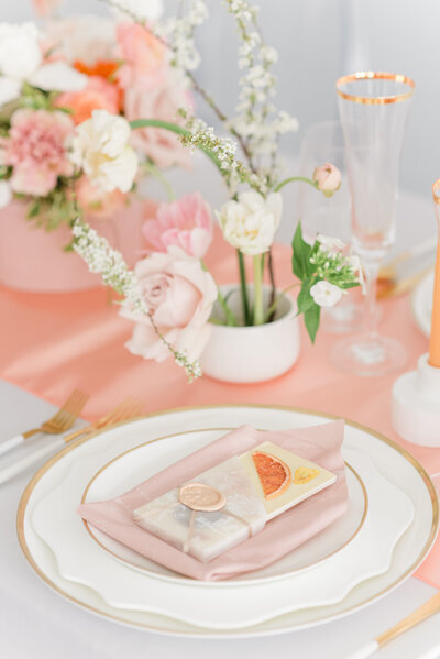 A table setting curated by a Banff wedding planner features a white and gold plate, pink napkin, floral arrangement, champagne flutes, and a small gift wrapped in pink.